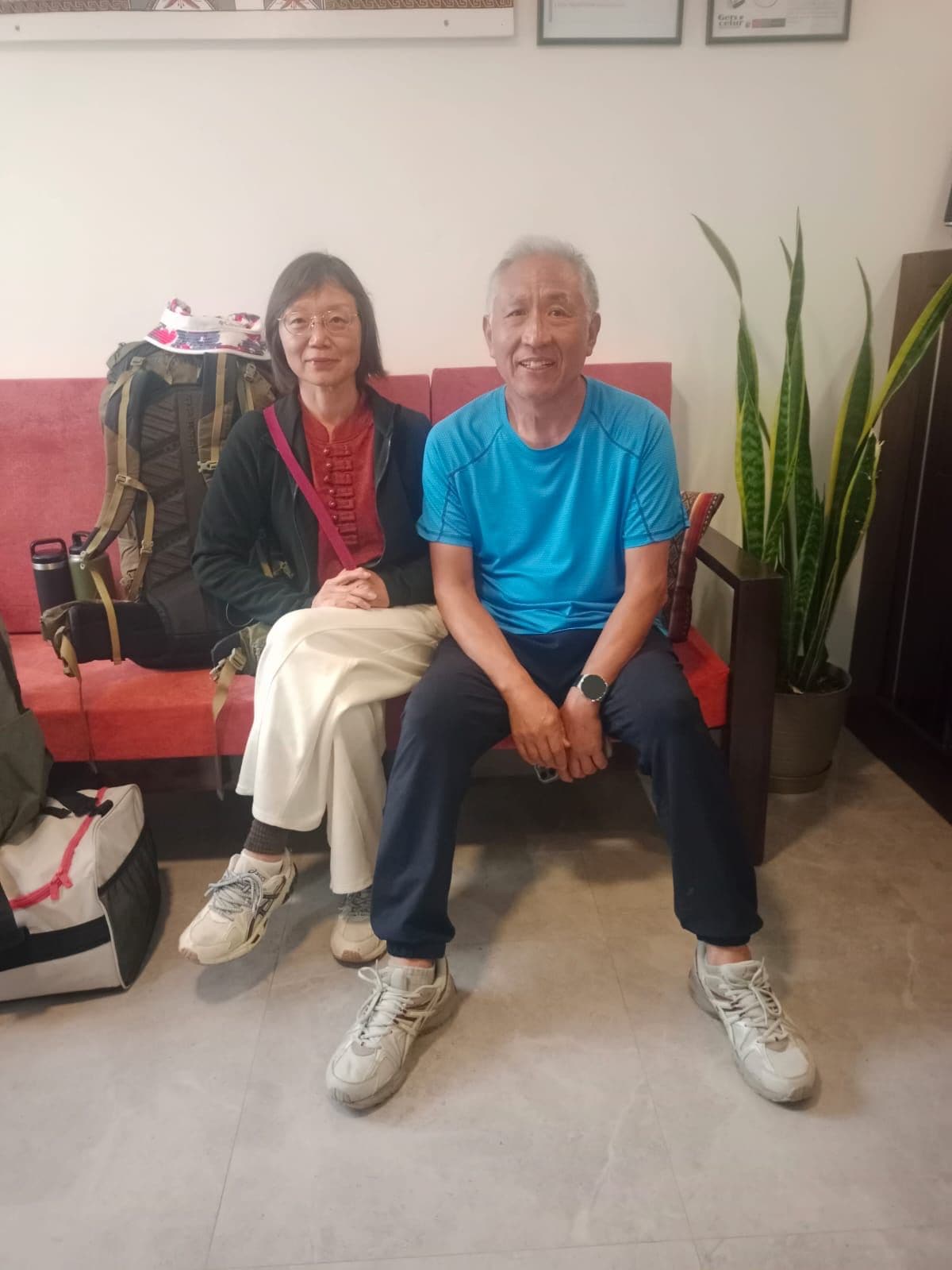 Two individuals seated on a red couch in a room with a tiled floor, a potted plant, and a framed picture on the wall.