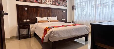 A hotel room with a large bed, a wooden headboard, a bedside table, a desk, and a large window with curtains.