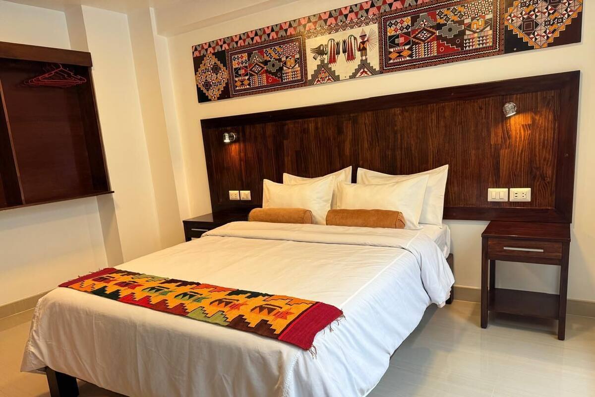 A hotel room with a large bed, a wooden headboard, two bedside tables, and a colorful wall hanging above the bed.