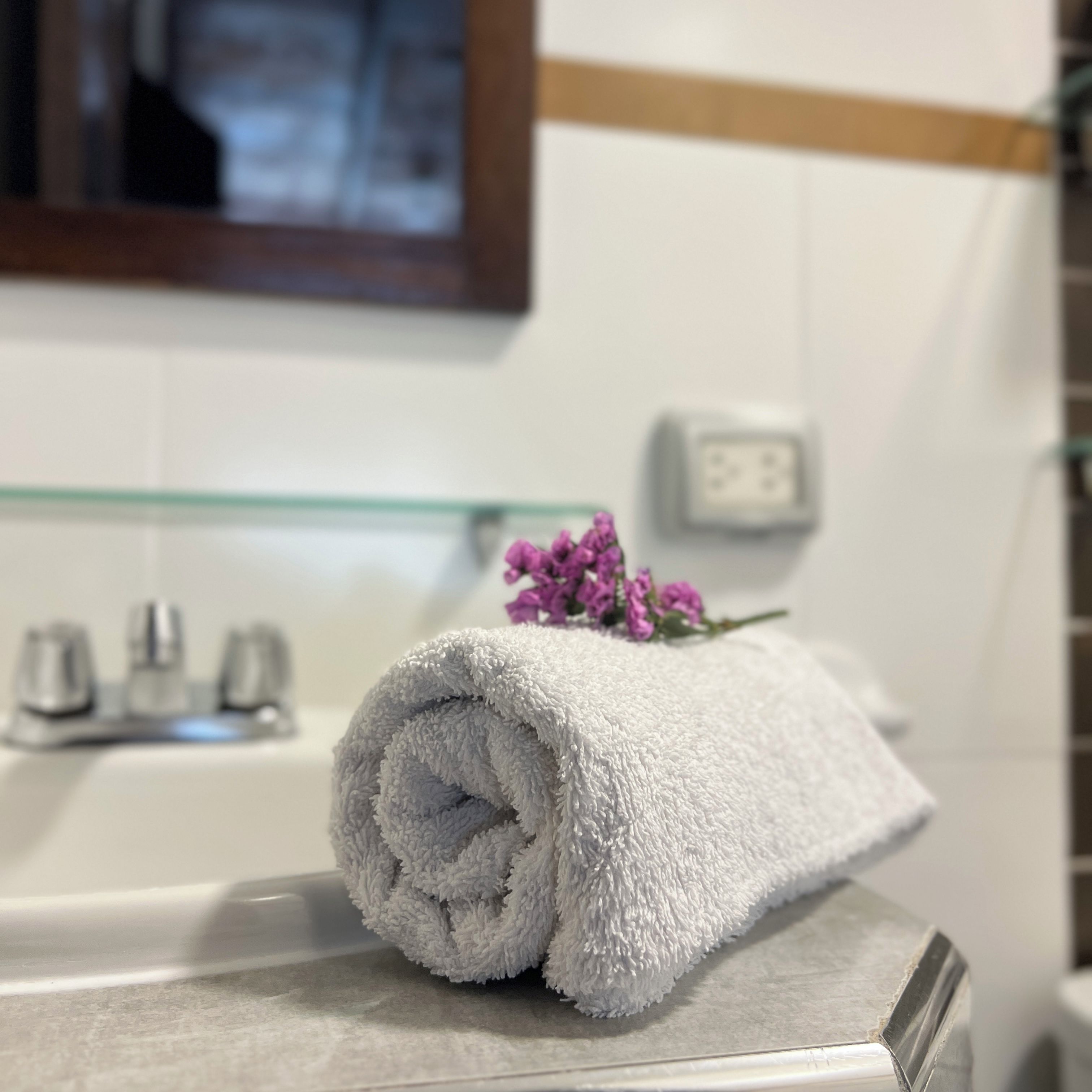 A bathroom with a white towel rolled up and a purple flower on top, a glass shelf, and a white wall with a switch.