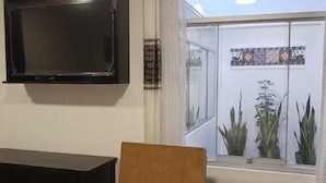 Un salon avec un téléviseur noir fixé au mur, un fauteuil marron, une table noire et une fenêtre donnant sur des plantes.