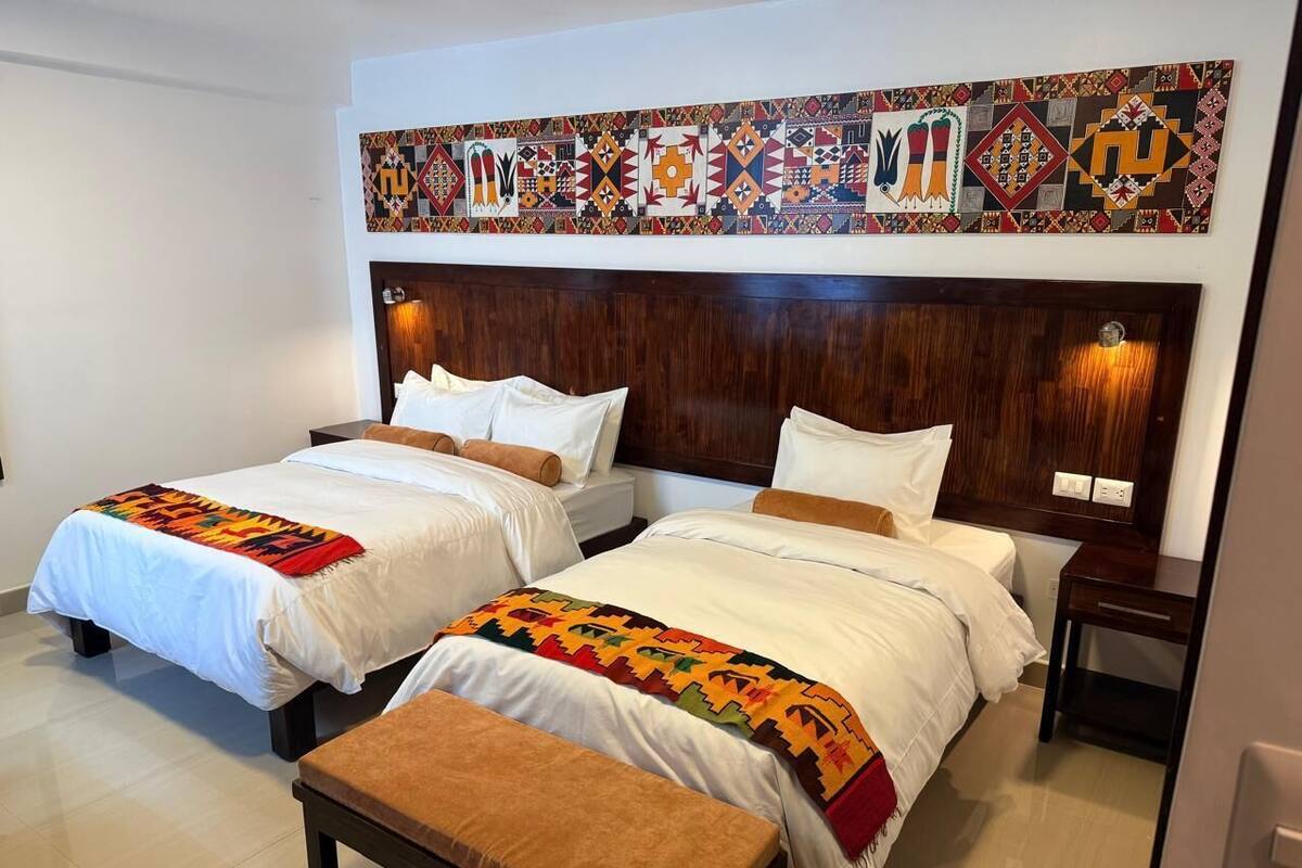 A hotel room with two beds, a wooden headboard, a colorful wall tapestry, and a small table.