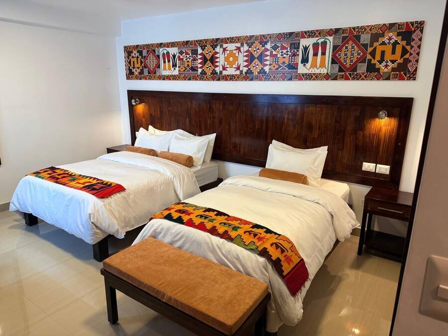 A hotel room with two beds, a wooden headboard, a colorful wall tapestry, and a small table.