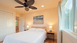 A bedroom with a bed, bedside lamps, a ceiling fan, a painting, and a window with curtains.