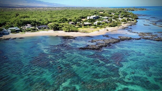 An aerial view of a coastal area with clear turquoise waters, a sandy beach, and a cluster of houses surrounded by greenery.
