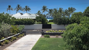 A modern house with a paved driveway leading to a gated entrance, surrounded by lush greenery and palm trees.