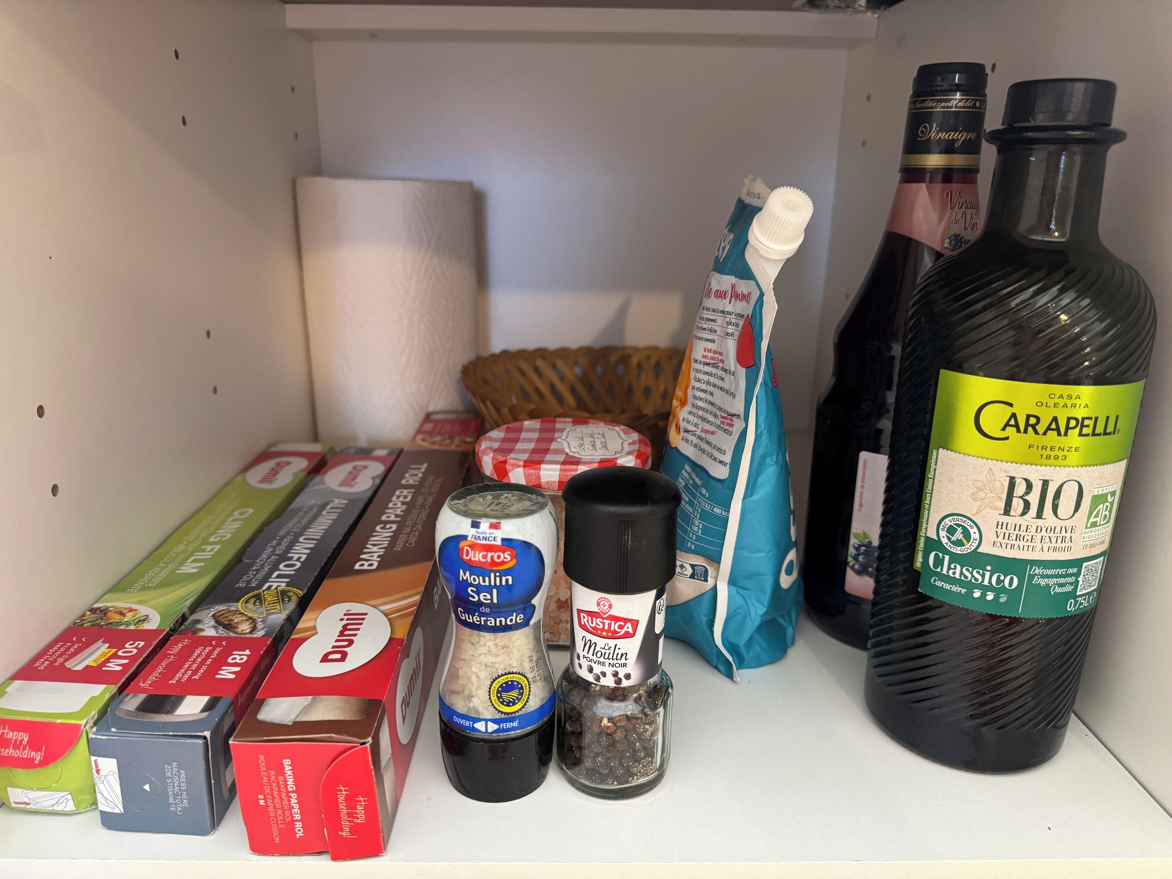 A shelf with various kitchen items including baking paper, flour, a bottle of olive oil, and a bag of flour.
