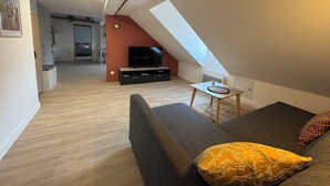 Un loft moderno con techo inclinado, un sofá con cojín estampado, una mesita y un televisor sobre un mueble negro.