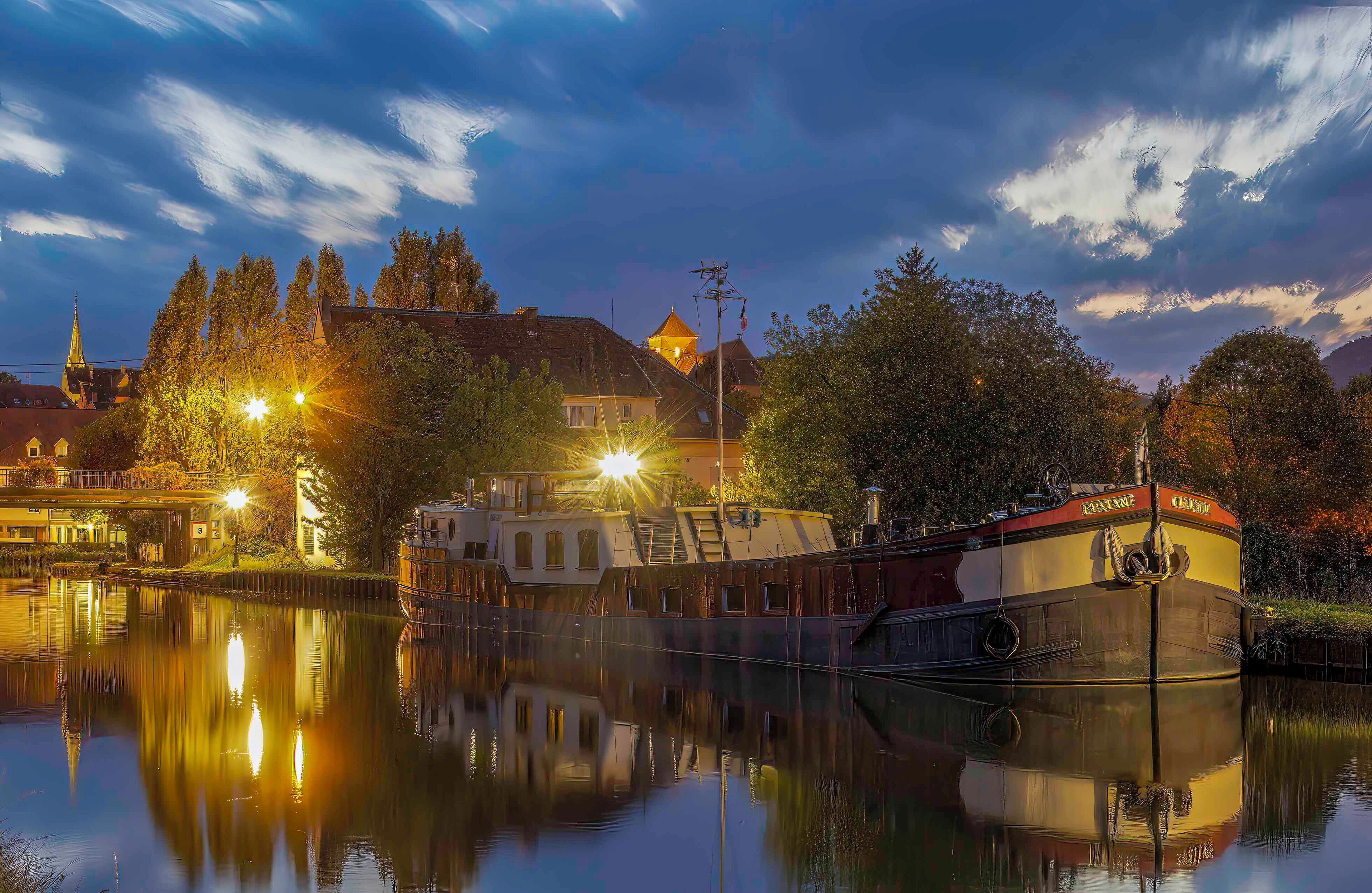 Une rivière paisible, avec un bateau amarré sur la rive droite, entourée d’arbres et de bâtiments dont les lumières se reflètent sur l’eau.