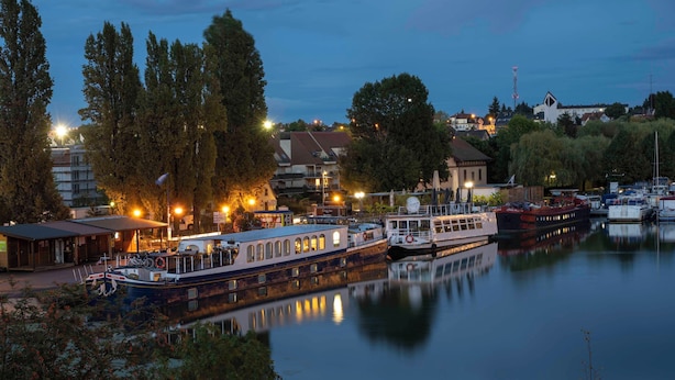 Un río tranquilo con barcos amarrados, rodeado de árboles y edificios con luces encendidas.