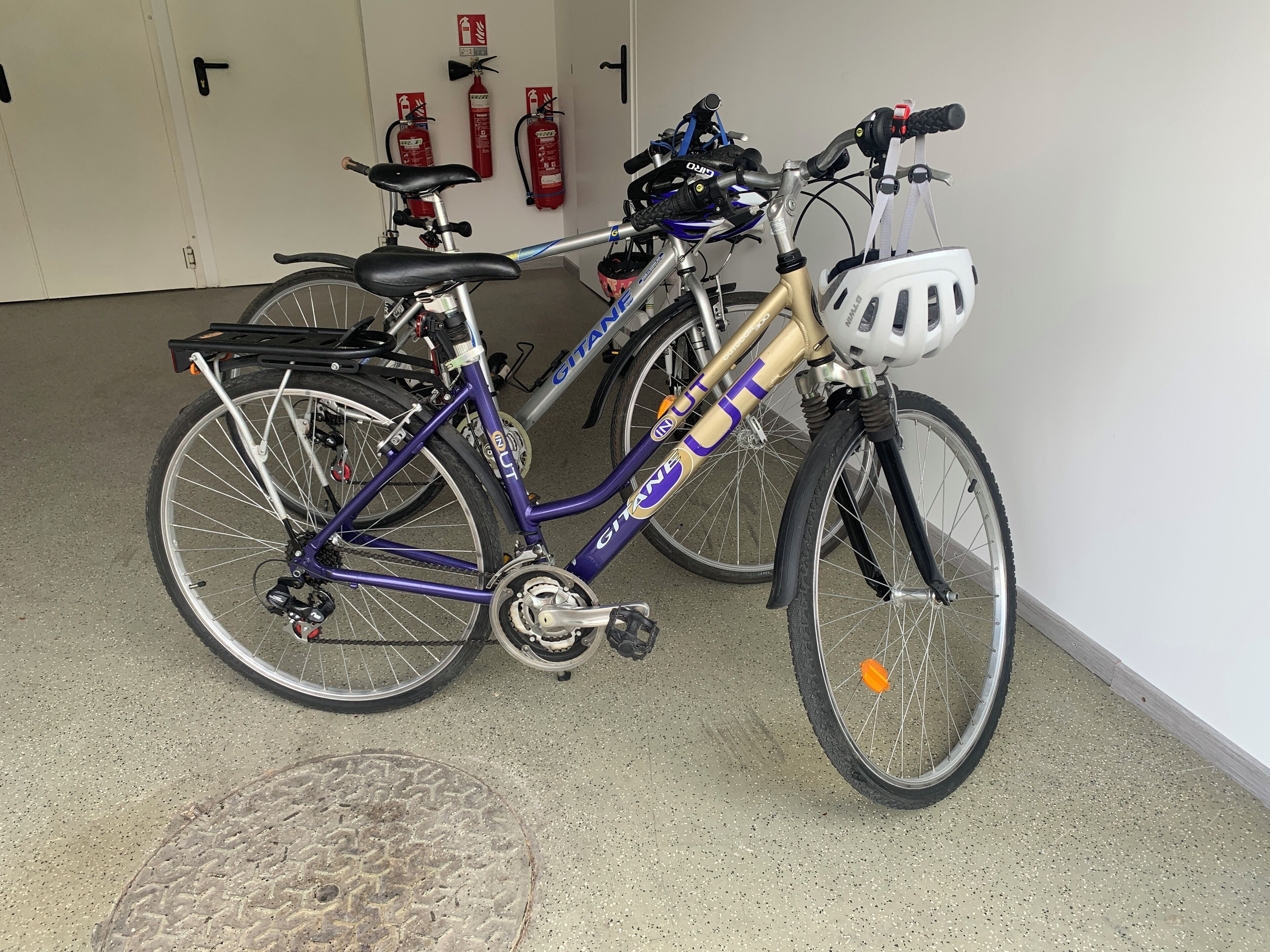 Een fiets met een wit mandje aan de voorkant en een gouden frame, geparkeerd binnen met brandblussers aan de muur.