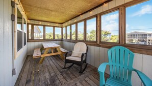 A covered porch with wooden flooring, a rocking chair, a turquoise chair, and a wooden table. - 4BR Hatteras Retreat • 5-Min Walk to Beach • Pet Friendly (Hatteras)