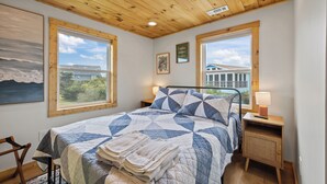 A bedroom with a bed, two windows, a wooden ceiling, and a view of a house. - 4BR Hatteras Retreat • 5-Min Walk to Beach • Pet Friendly (Hatteras)
