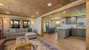 A spacious living room with a sofa, coffee table, and rug, leading to a kitchen with grey cabinets and a dining area. - 4BR Hatteras Retreat • 5-Min Walk to Beach • Pet Friendly (Hatteras)