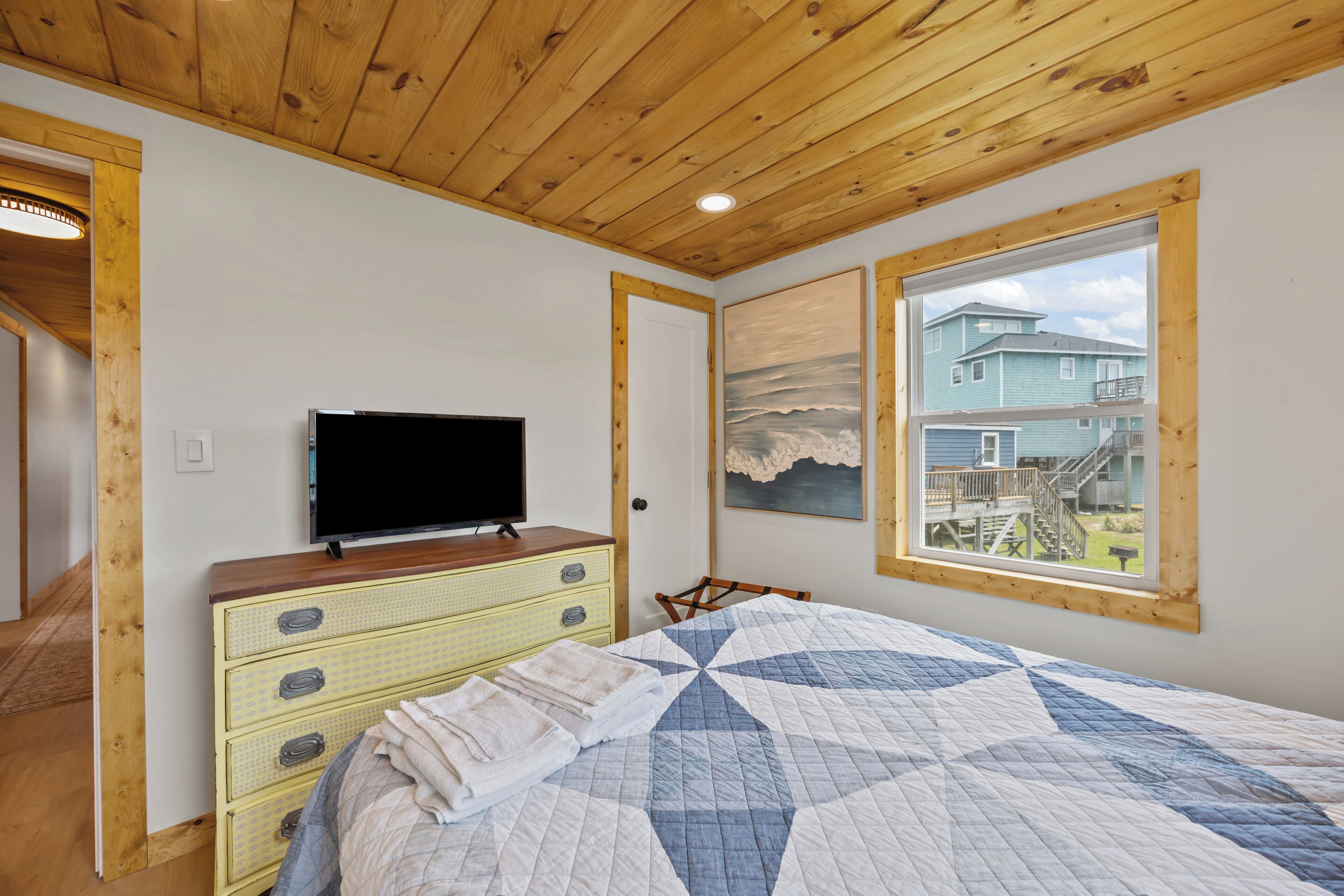 A bedroom with a wooden ceiling, a bed with a quilt, a dresser, a television, and a window with a view of a house.