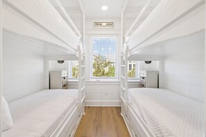 A room with bunk beds, a window with blinds, a sign above the window, and a small table with a lamp.