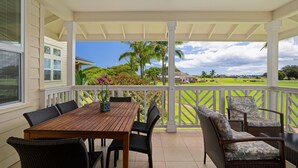 A covered patio with a wooden table and wicker chairs, overlooking a green landscape with palm trees.