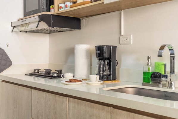 Una cocina con estufa a gas, cafetera, pileta y una alacena con utensilios de cocina.