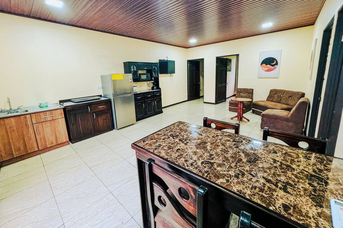 A spacious living area with a kitchen, dark wooden cabinets, a granite countertop, and a television mounted on the wall.