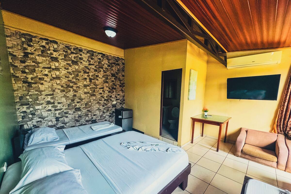 A room with a bed, a small table, a chair, a television, and a stone wall.