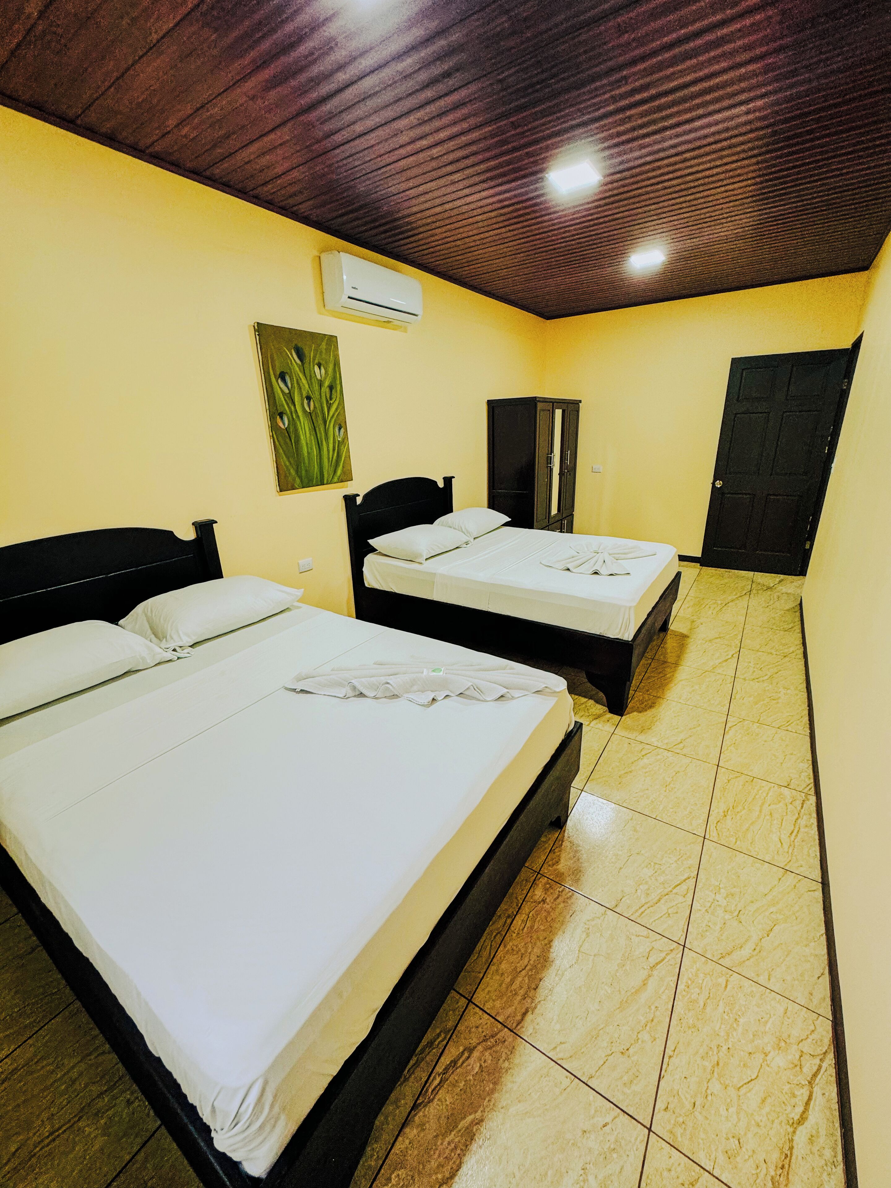 A hotel room with two beds, a wardrobe, a painting, and an air conditioning unit.