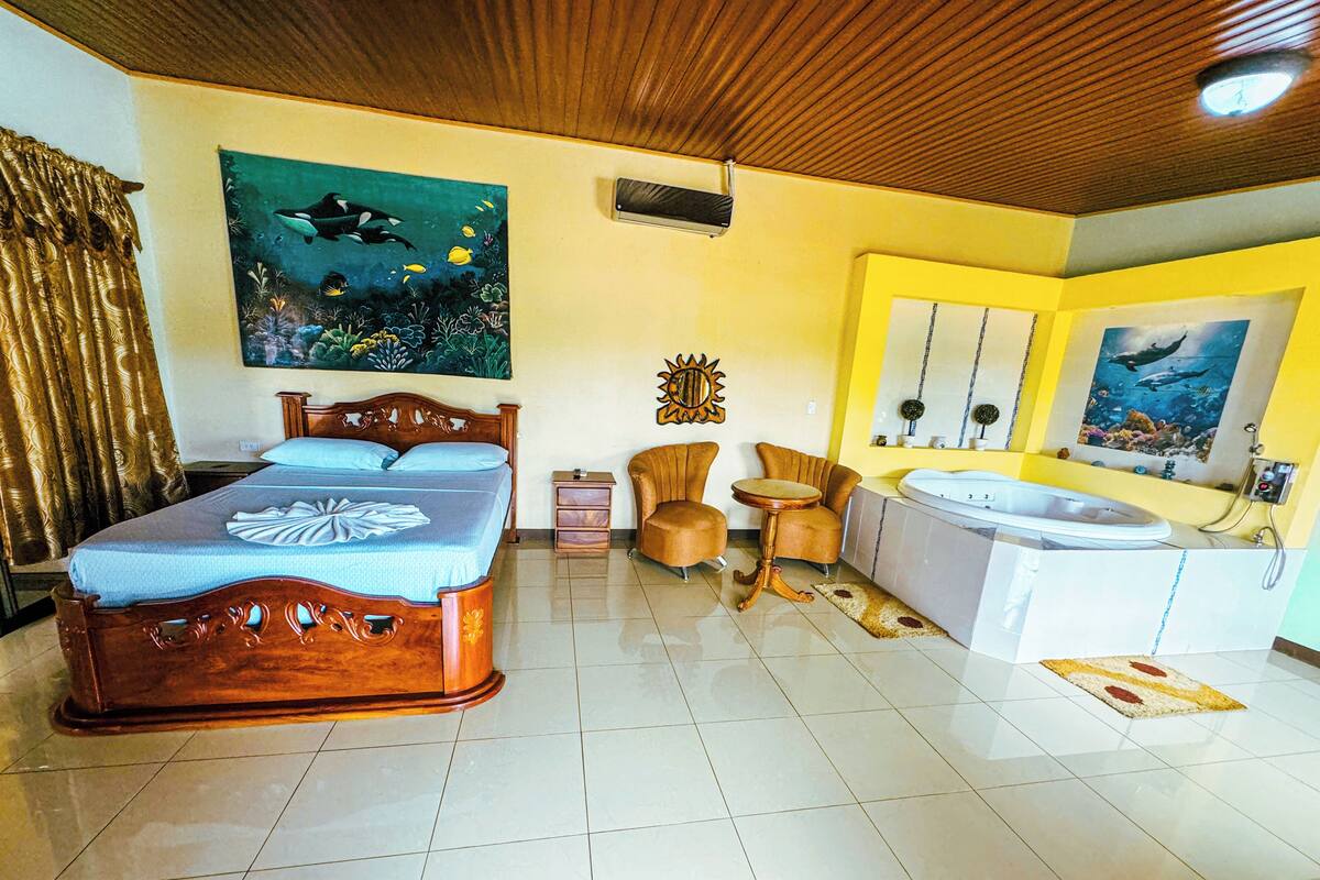 A hotel room with a bed, a small table, two armchairs, a sofa, a bathtub, and a large window.