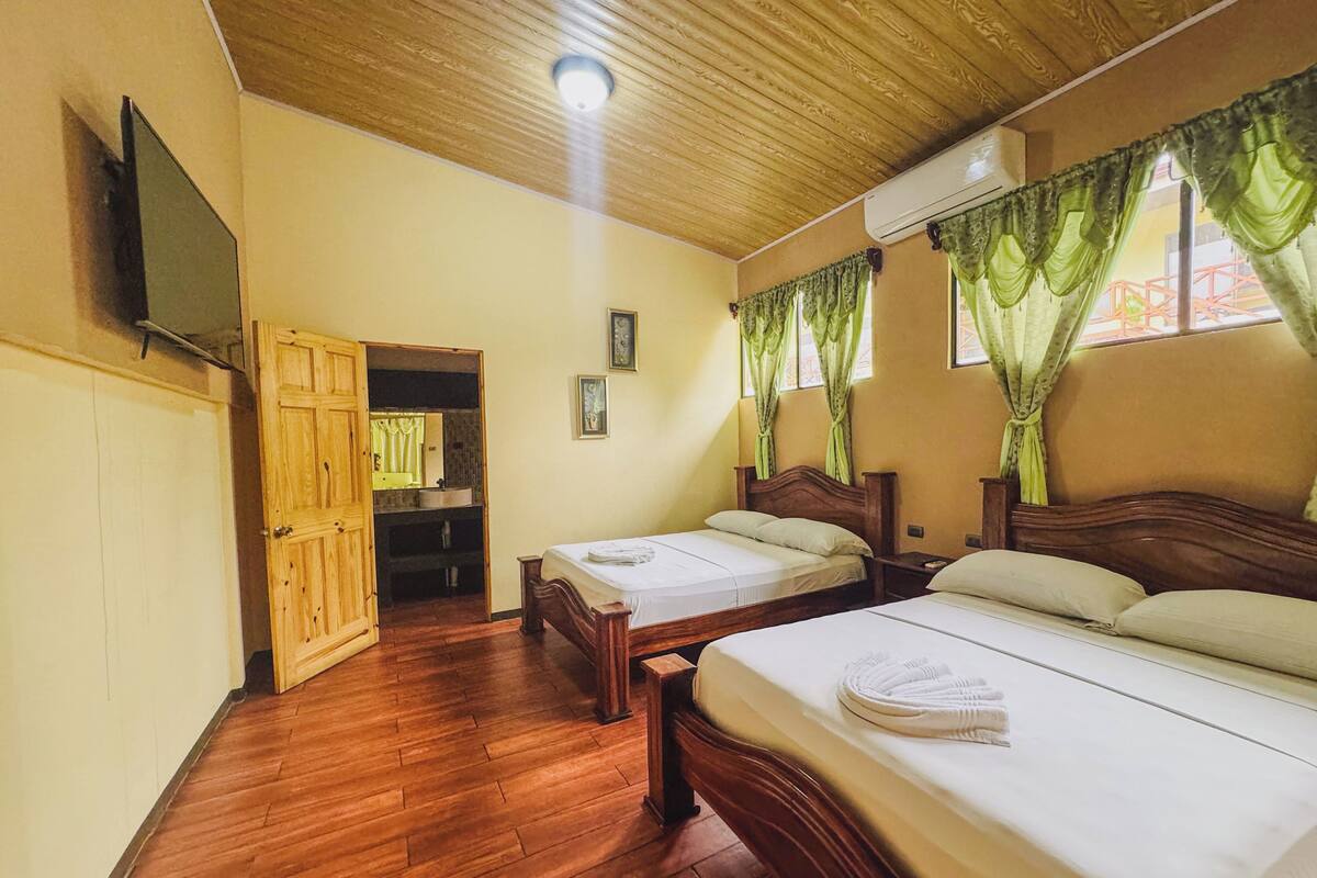 A hotel room with two wooden beds, a wooden ceiling, a flat-screen TV mounted on the wall, and a bathroom visible through an open door.