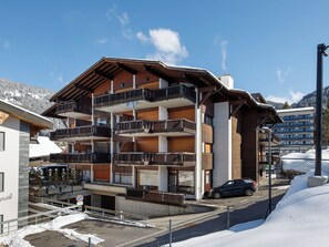 A multi-story hotel with balconies, a parking area, and a snowy mountain backdrop.