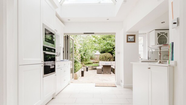 A bright kitchen with white cabinets, a built-in oven, and a glass door leading to a garden patio with outdoor furniture. - Stylish Townhouse Annexe Cheltenham (Cheltenham)