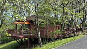 A rustic wooden cabin with a balcony, surrounded by trees and a gravel path.