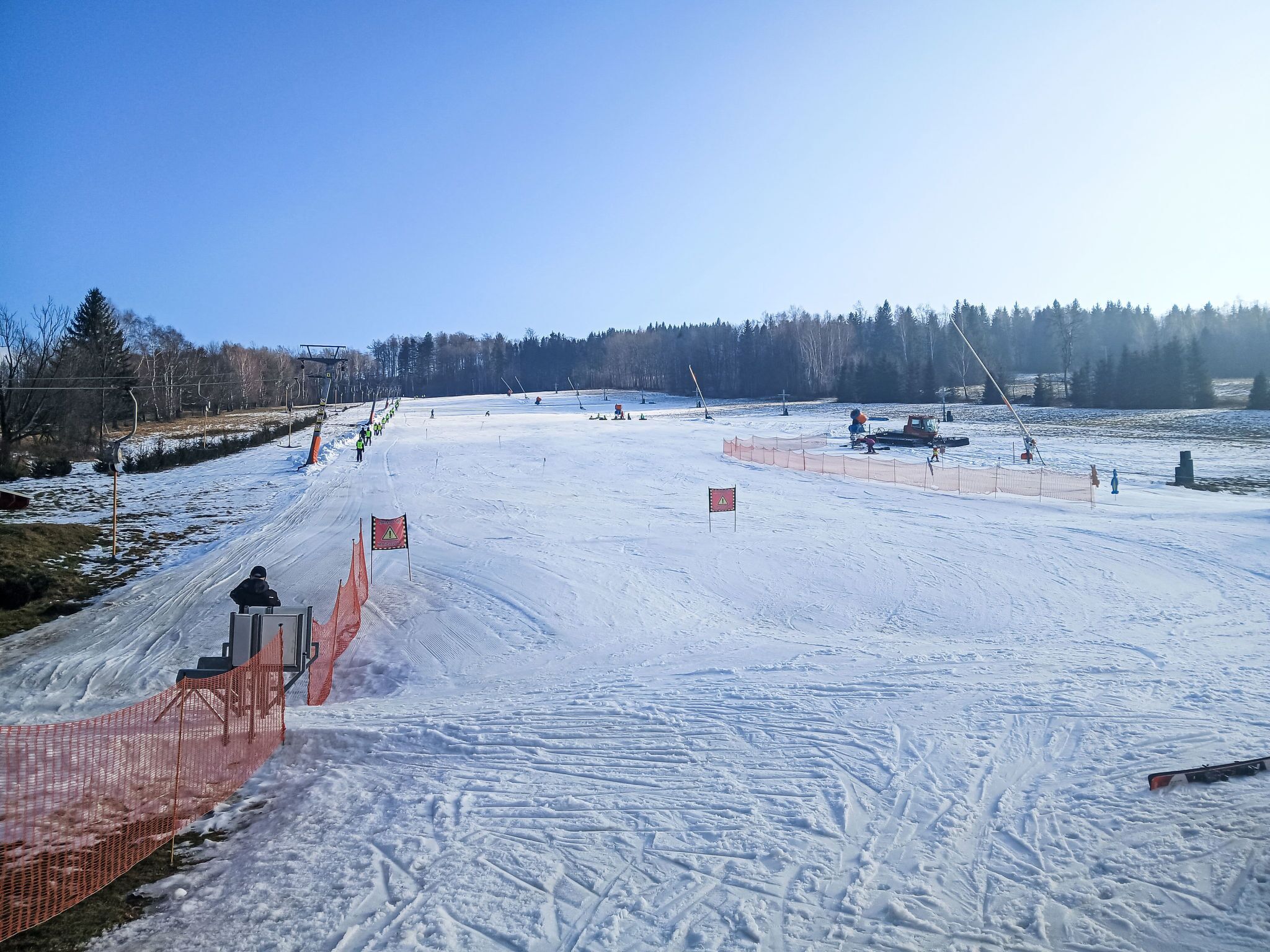 A snowy ski slope with orange safety nets and flags, surrounded by trees and a clear sky.