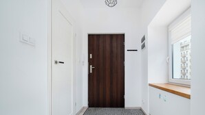 A narrow hallway with a wooden door, a white wall, a window with a view of buildings, and a small wooden counter.
