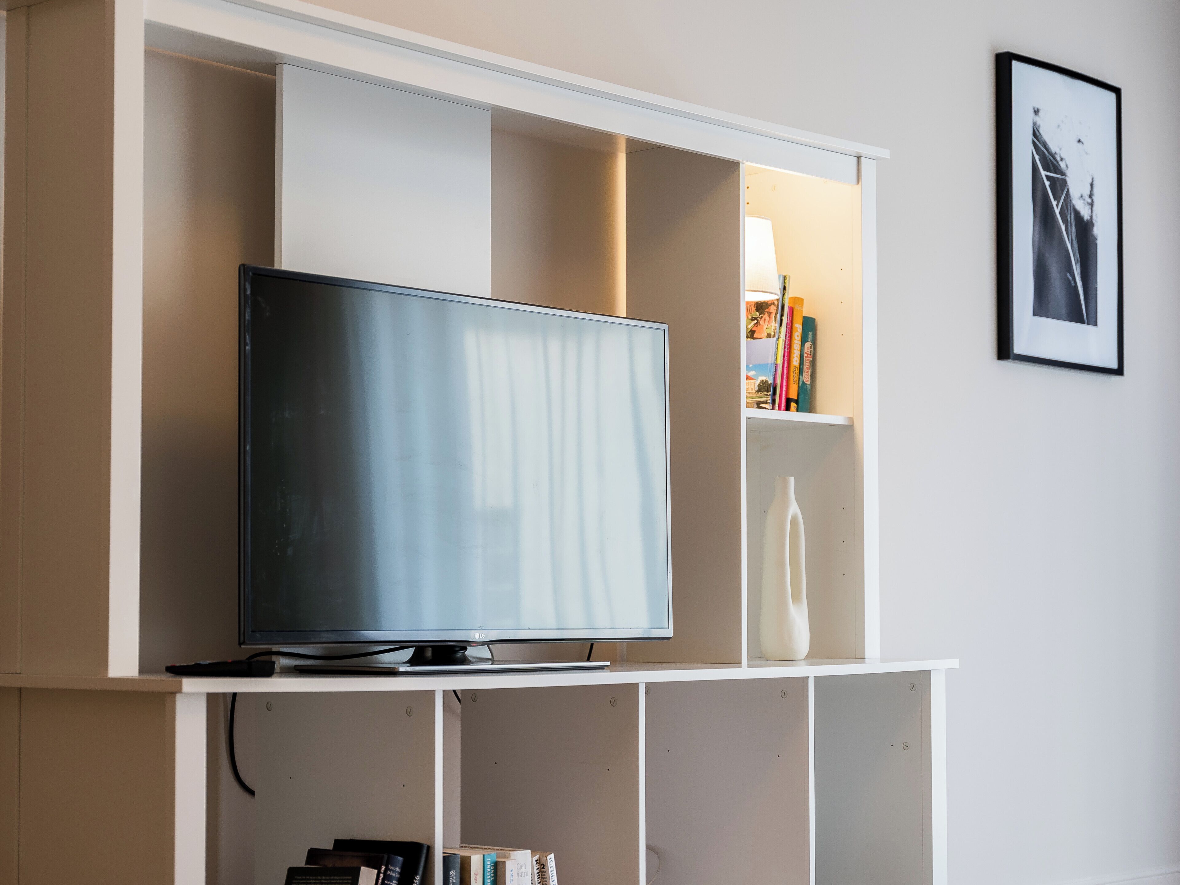 A modern living room with a flat-screen TV on a white stand, a bookshelf with books and decorative items, and a framed picture on the wall.