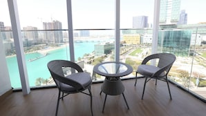 Un balcon avec deux fauteuils en osier et une table en verre, offrant une vue sur le paysage urbain et une étendue d’eau.
