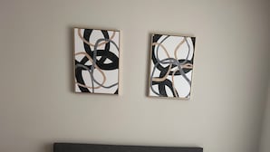 Un lit bien fait, avec une literie blanche et une tête de lit grise, encadré de deux tableaux abstraits accrochés au mur.