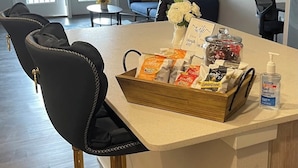 Un coin bar avec un tabouret, un plateau de collations, un distributeur de gel hydroalcoolique et un vase à fleurs.