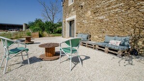 Een terras met een stenen muur, twee rieten stoelen, een houten tafel en een bank met kussens.