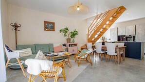 Een moderne woonkamer met een groene bank, rieten stoelen, een eettafel en een keuken met een houten trap.