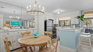A modern kitchen with white cabinetry, a central island, and a dining area with wooden chairs and a round table. - Elegant Beach Condo-Views of the beach, pool, and courtyard right from balcony!! (Destin)