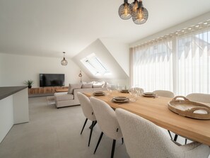 Un espace de repas moderne et bien éclairé, avec une table en bois, des chaises blanches et un téléviseur en arrière-plan.