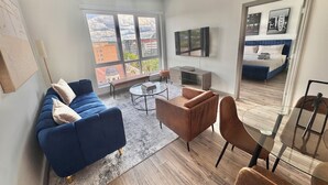 Uma sala de estar moderna com um sofá azul, uma poltrona marrom e uma mesa de centro de vidro. Uma televisão está instalada na parede, e há vista para um horizonte urbano por grandes janelas.