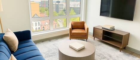 Uma sala de estar moderna com um sofá azul, uma mesa de centro redonda e vista para edifícios através de amplas janelas.