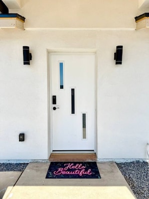 Una puerta blanca con panel vertical y horizontal, un felpudo con la inscripción 'Hello Beautiful', y dos luces fijadas a la pared.