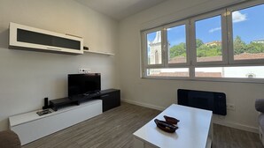 Un salon moderne doté d’un téléviseur à écran plat, d’une table blanche et d’une grande fenêtre donnant sur un immeuble et des arbres.