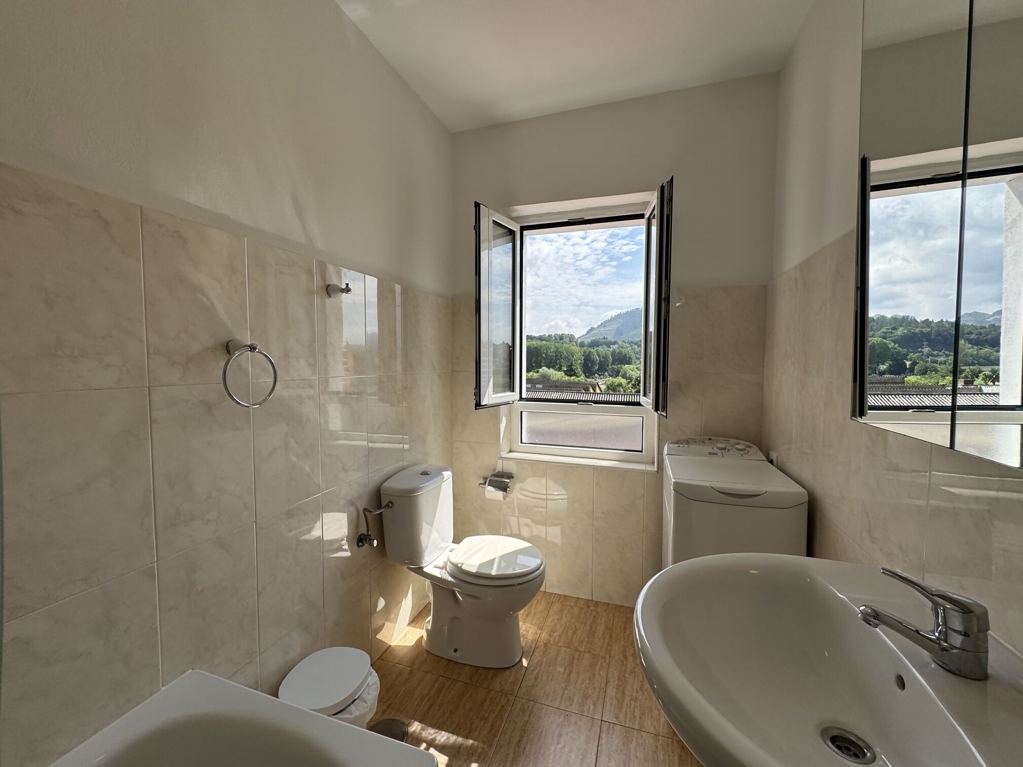 A modern bathroom with a toilet, bidet, and bathtub. A window provides a view of a green landscape.