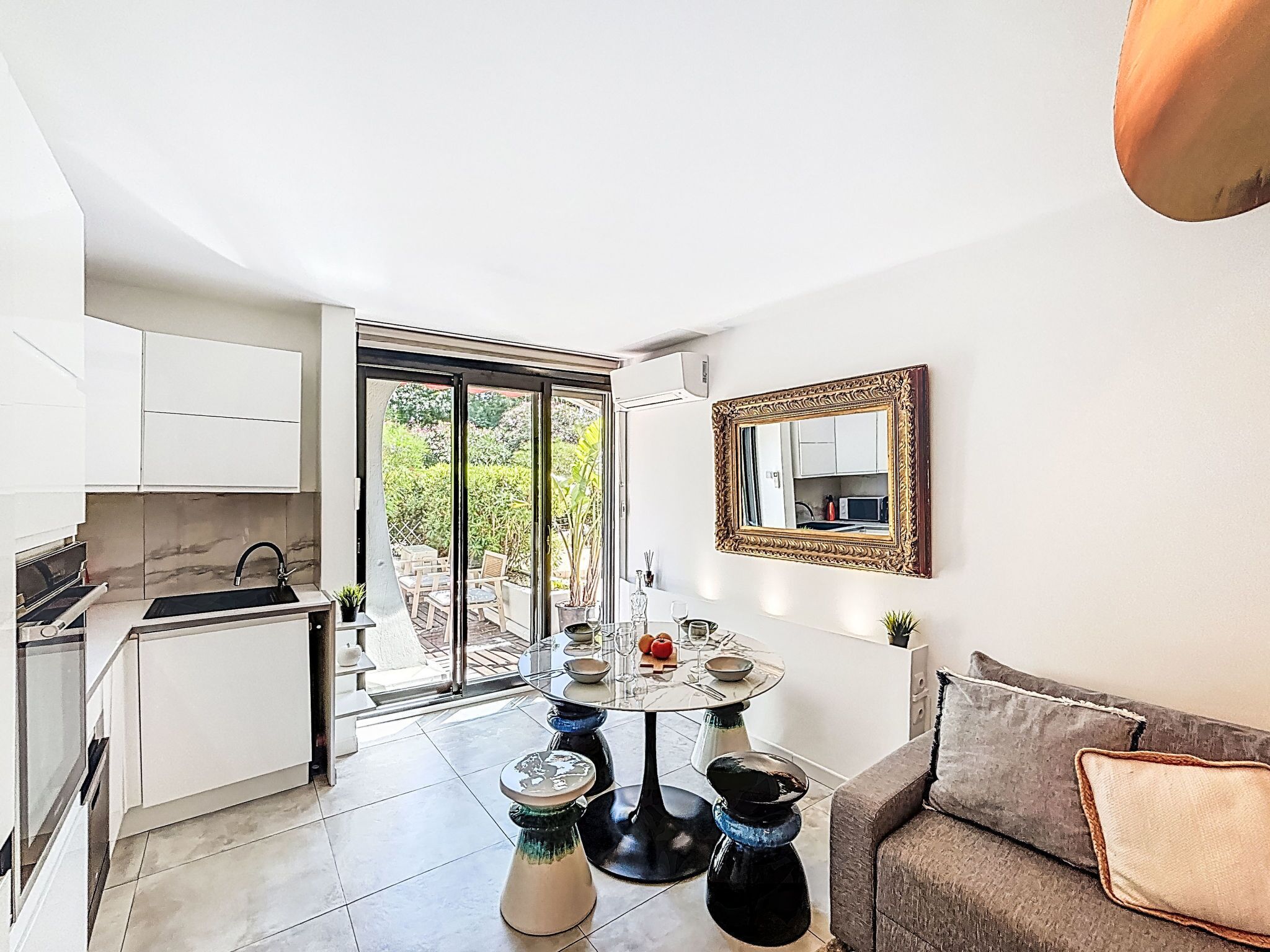 A modern kitchen with white cabinets, a dining area with a round table and stools, a sofa with cushions, and a large mirror on the wall.