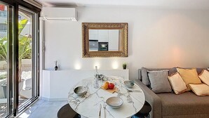 Uma sala de estar moderna com uma mesa redonda de mármore posta para refeições, um sofá com almofadas, um espelho grande na parede e uma porta de vidro deslizante que dá para uma área externa.