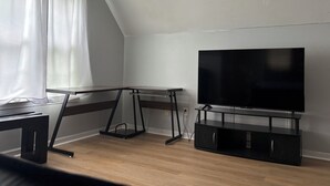 Et værelse med skråvæg, et stort fladskærms-tv på et sort stativ, et træbord og et vindue med hvide gardiner.