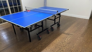 Une table de ping-pong bleue, munie de roulettes, posée sur un plancher en bois.