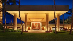 Una entrada de hotel moderna con una gran obra de arte iluminada, palmeras y un césped cuidado.